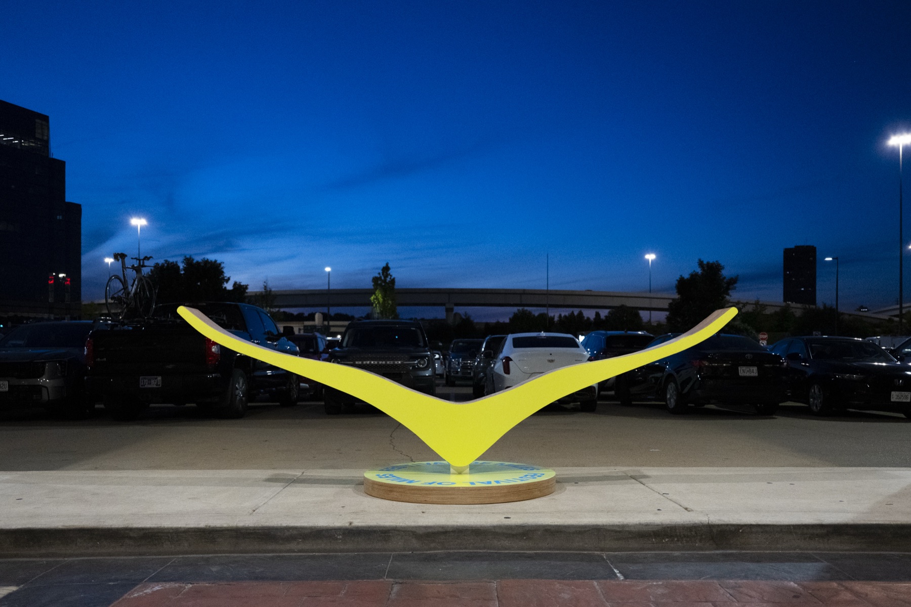 Sculpted plywood HOKA wing in campaign yellow on a printed Festival of Miles plinth at dusk