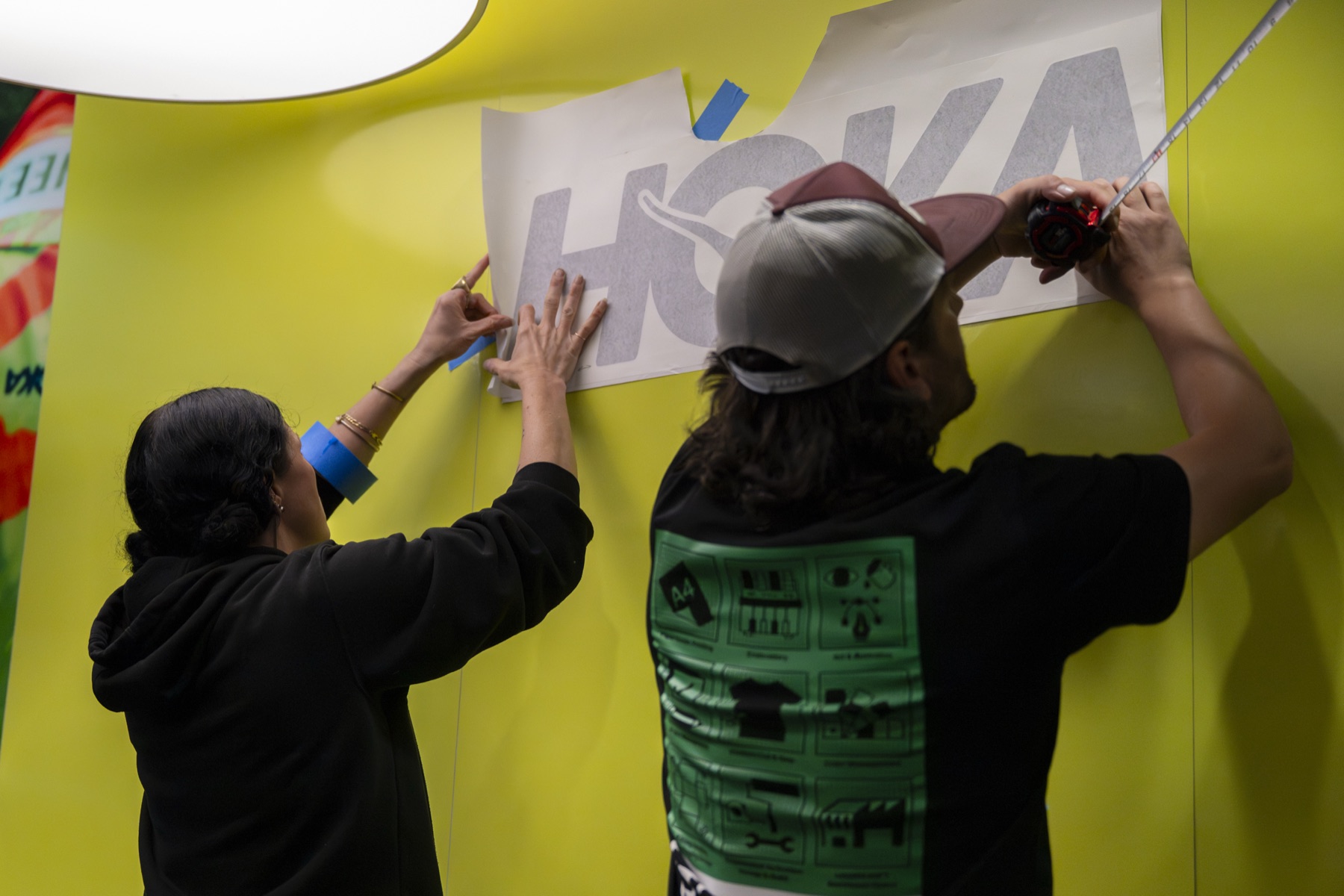 Two Fieldkit installers applying contour-cut HOKA logotype vinyl to a yellow campaign wall at Festival of Miles